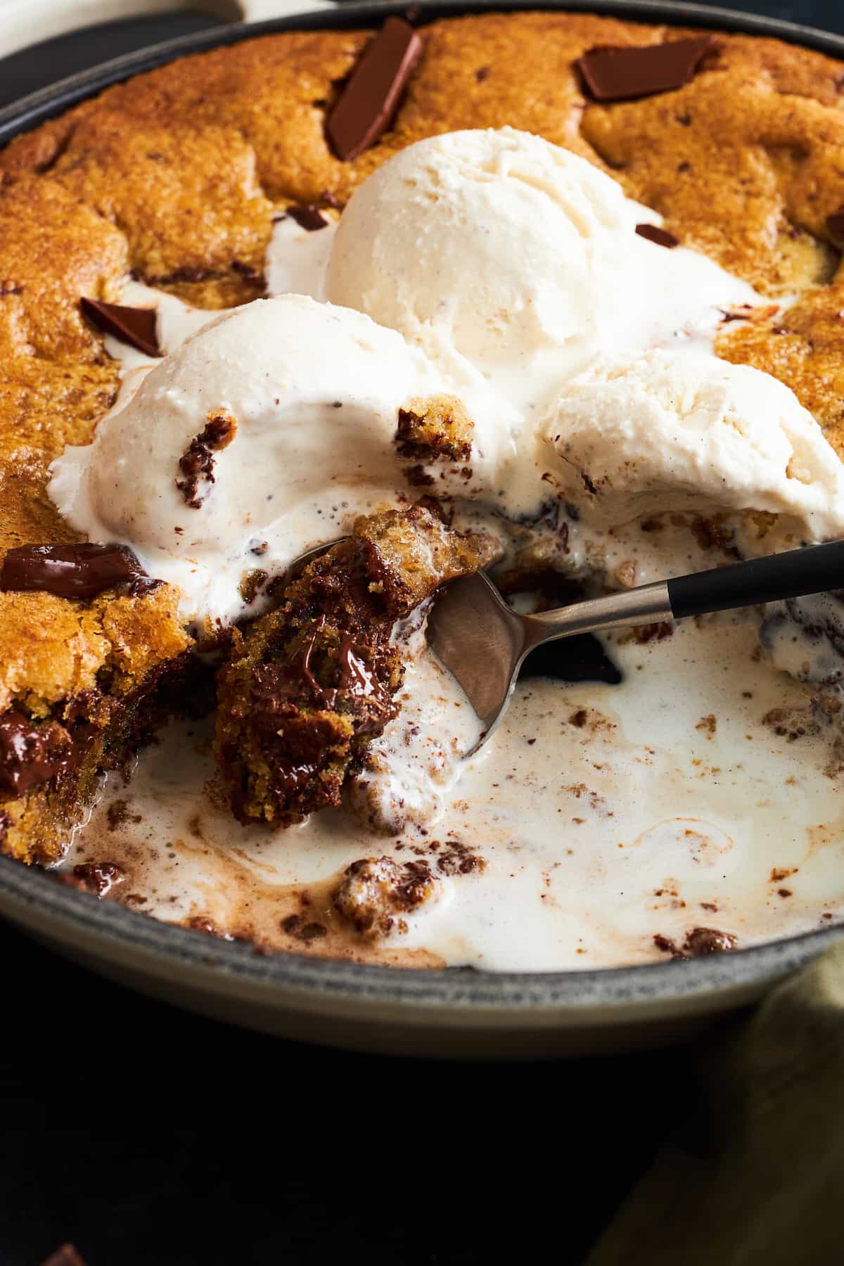 A spoon digging into a skillet chocolate chip cookie topped with vanilla ice cream. 