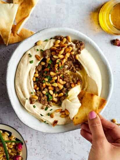 A bowl of hummus with ground beef with a pita chip being dipped in.