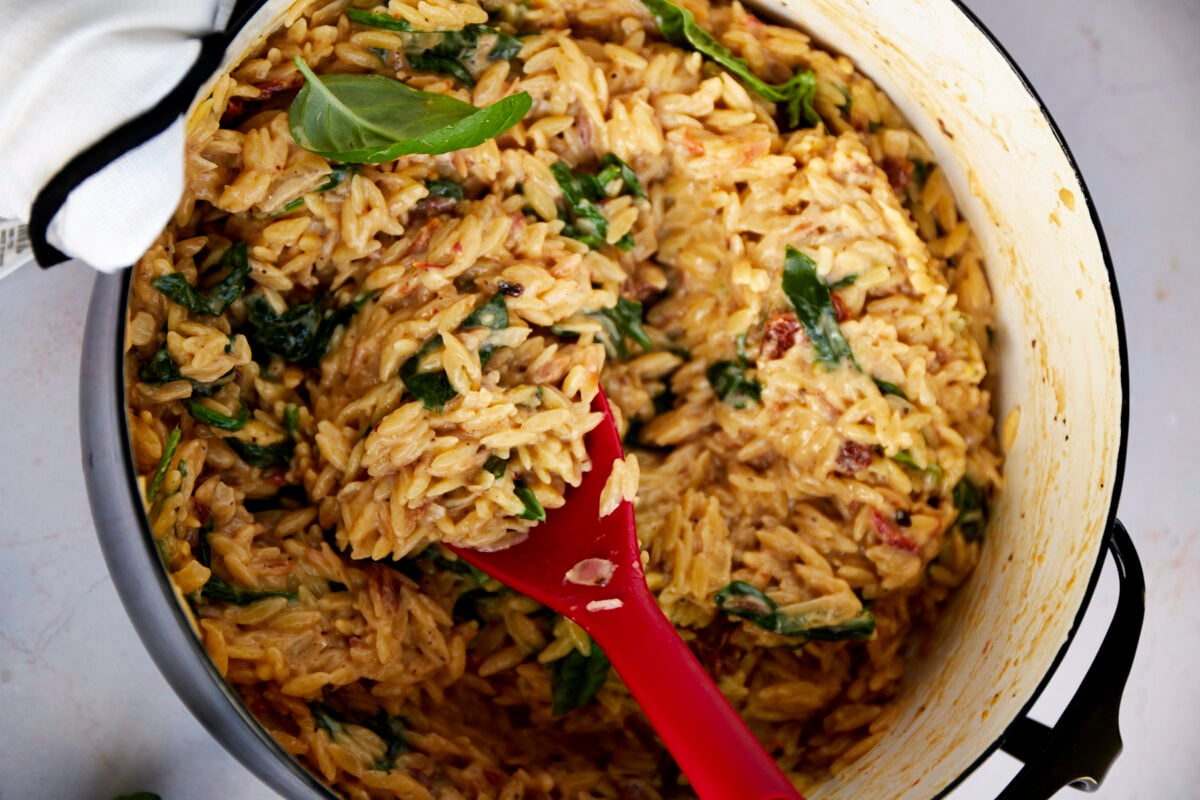 Creamy Tuscan orzo in a pot, a red spoon lifting a scoop. 