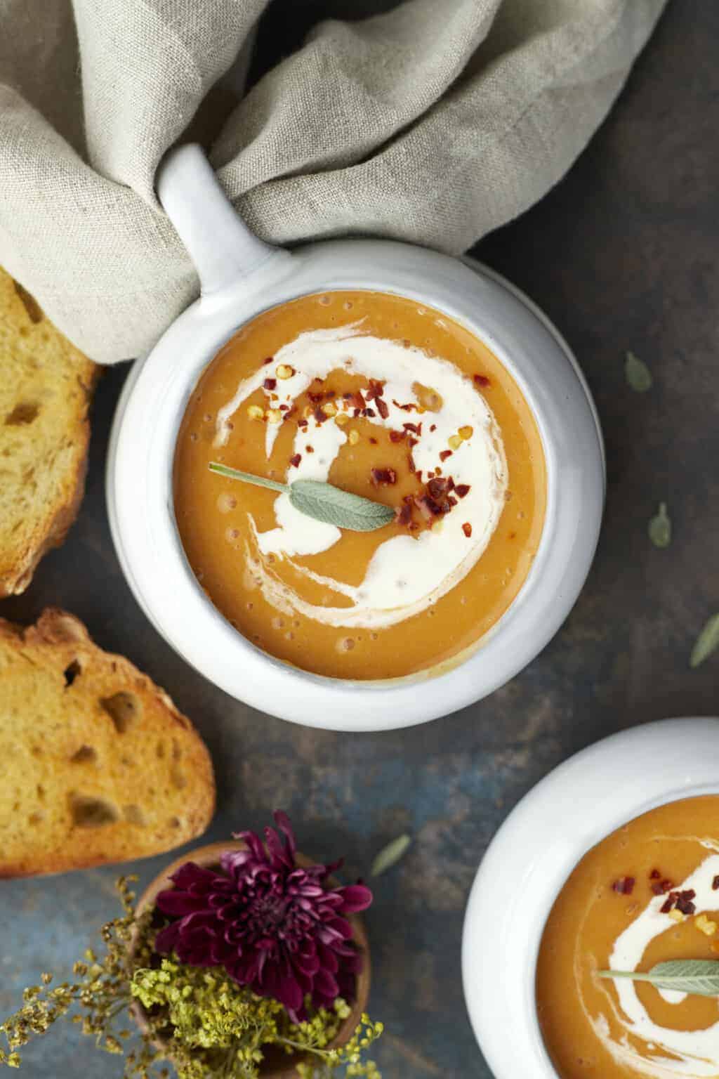 A bowl of creamy orange soup garnished with a swirl of cream, crumbled red pepper, and a sage leaf, with slices of toasted bread and a purple flower nearby.