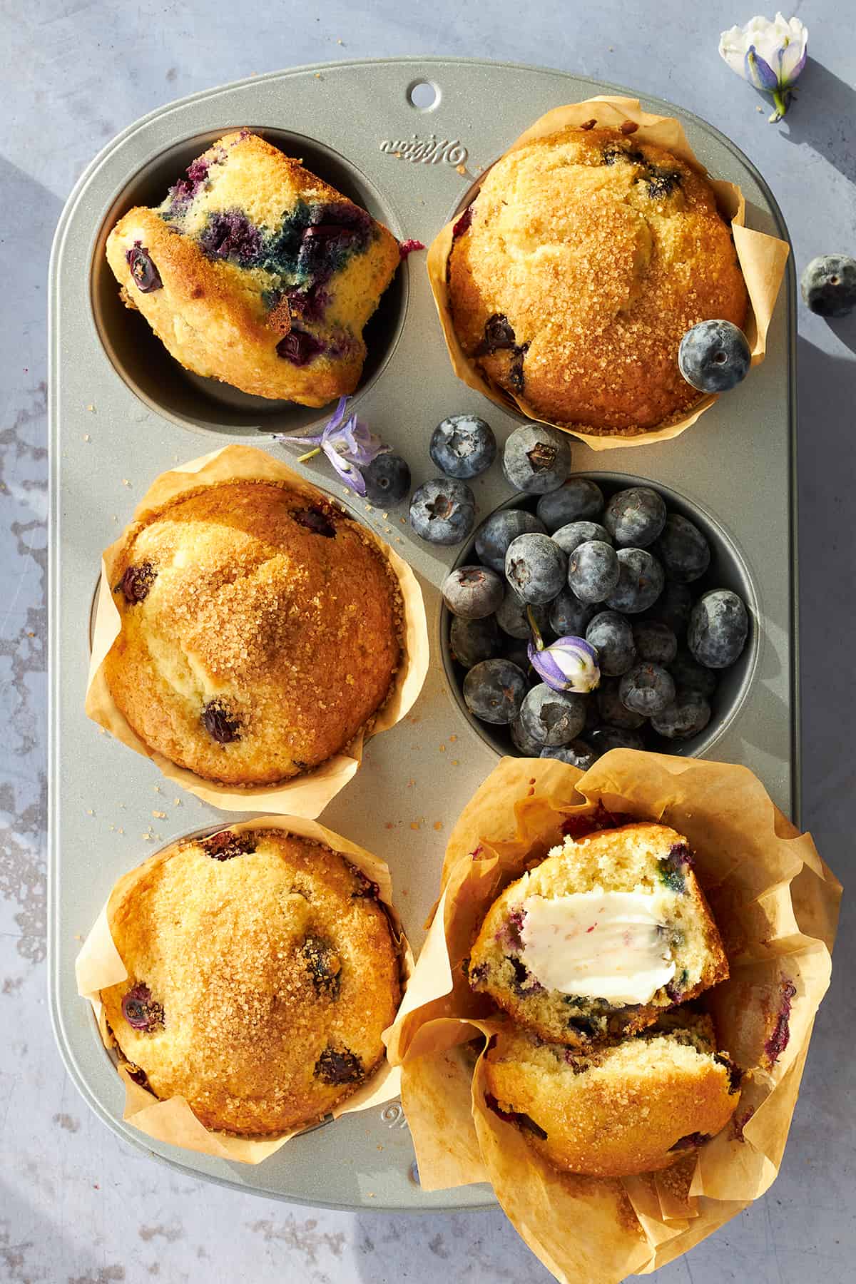 Baked blueberry muffins in a muffin tin, one sliced and topped with butter.