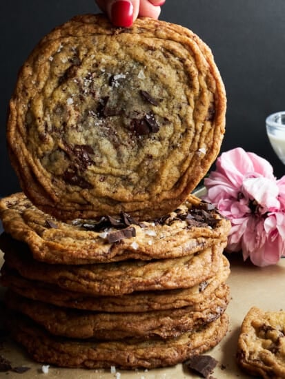 A hand lifting a banging chocolate chip cookie off a stack of cookies.