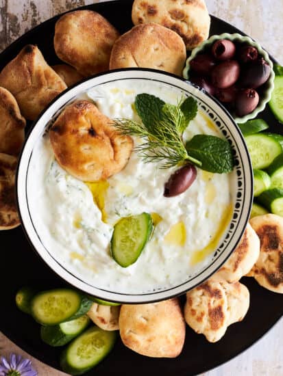 A bowl of tzatziki sauce on a platter with pita bread, olives, and cucumbers.