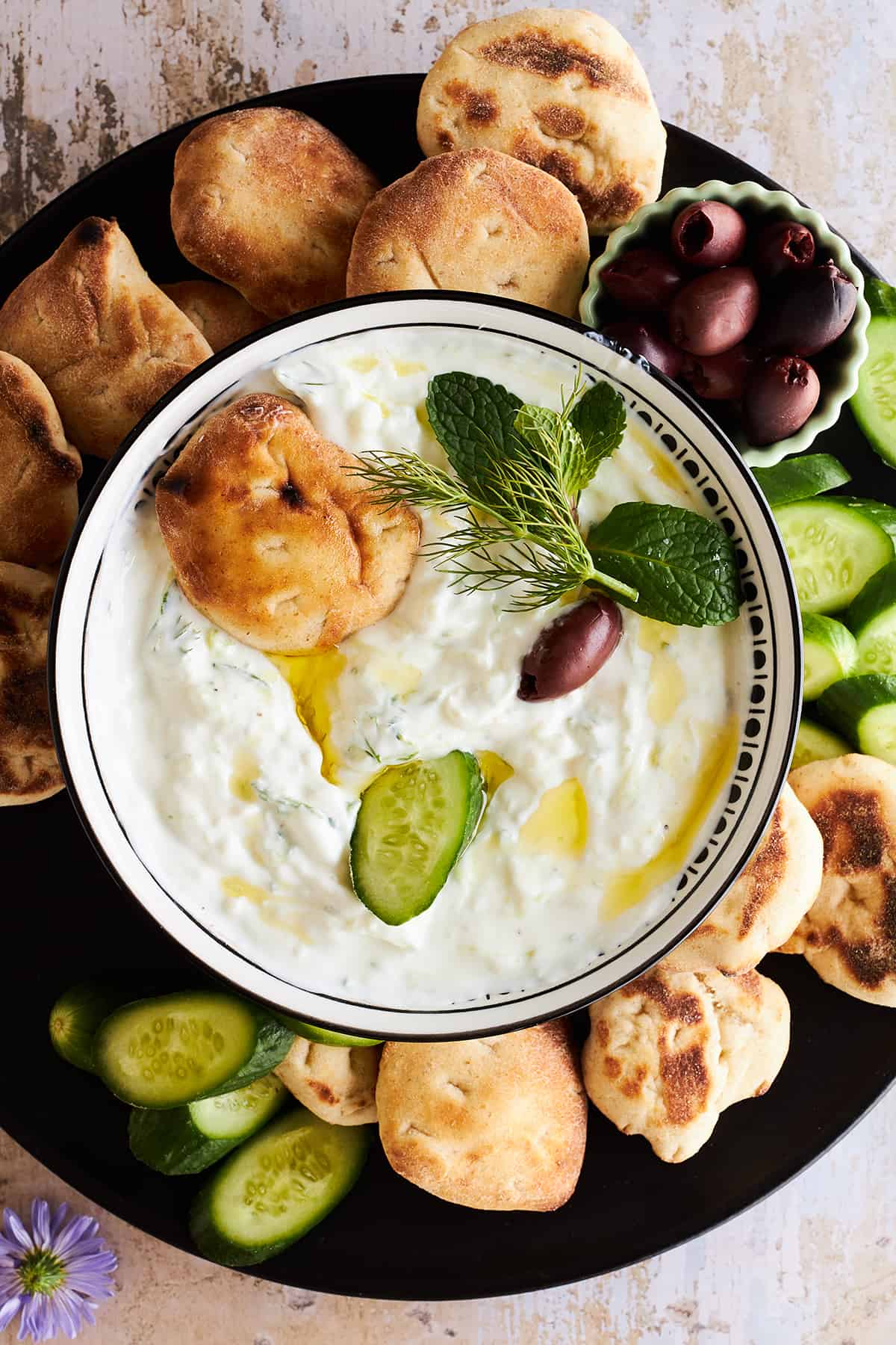 A bowl of tzatziki sauce on a plate with pita, olives, and cucumber slices. 