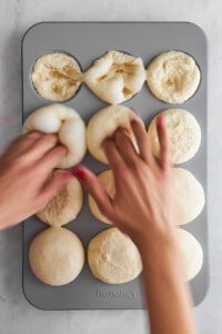 Two hands making indents in raw dinner rolls in a muffin tin.