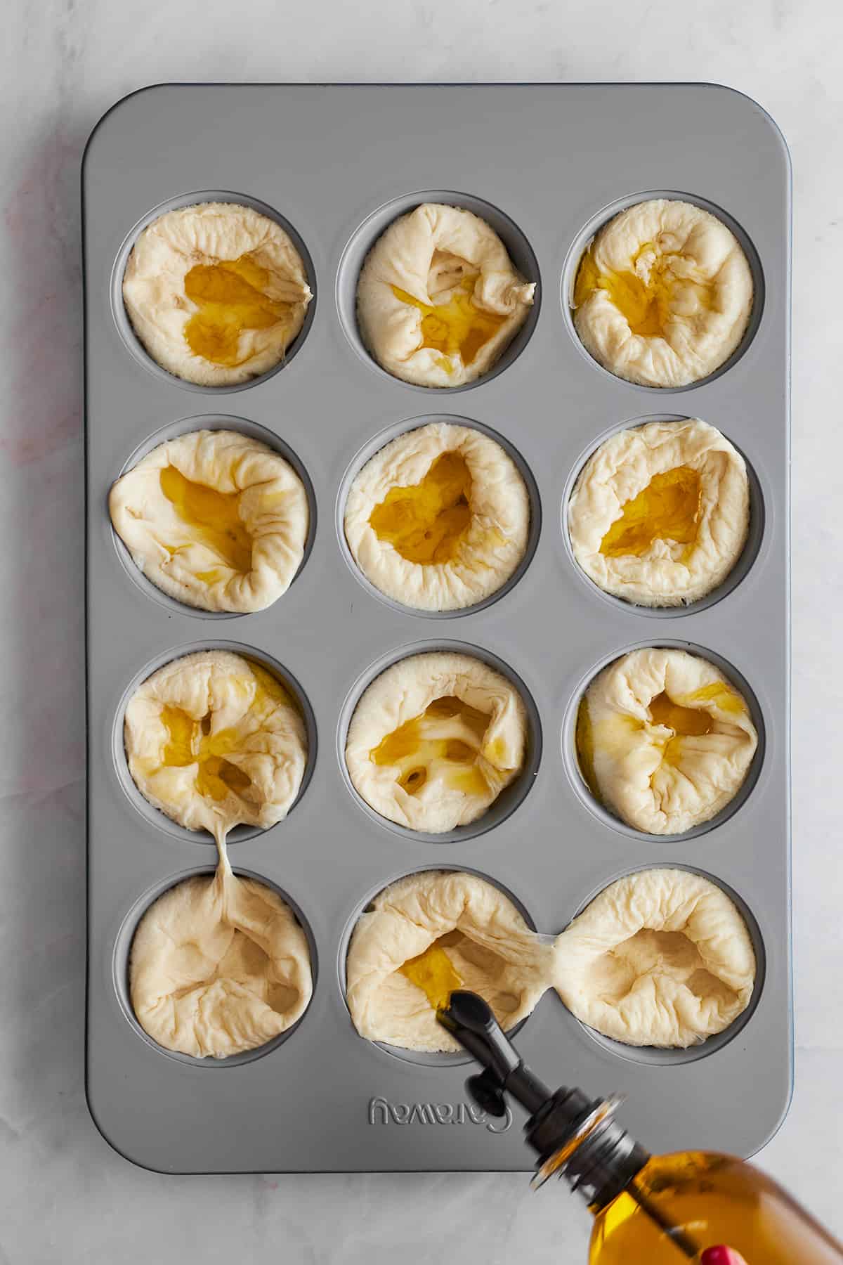 Olive oil being drizzled over bread rolls in a muffin tin. 