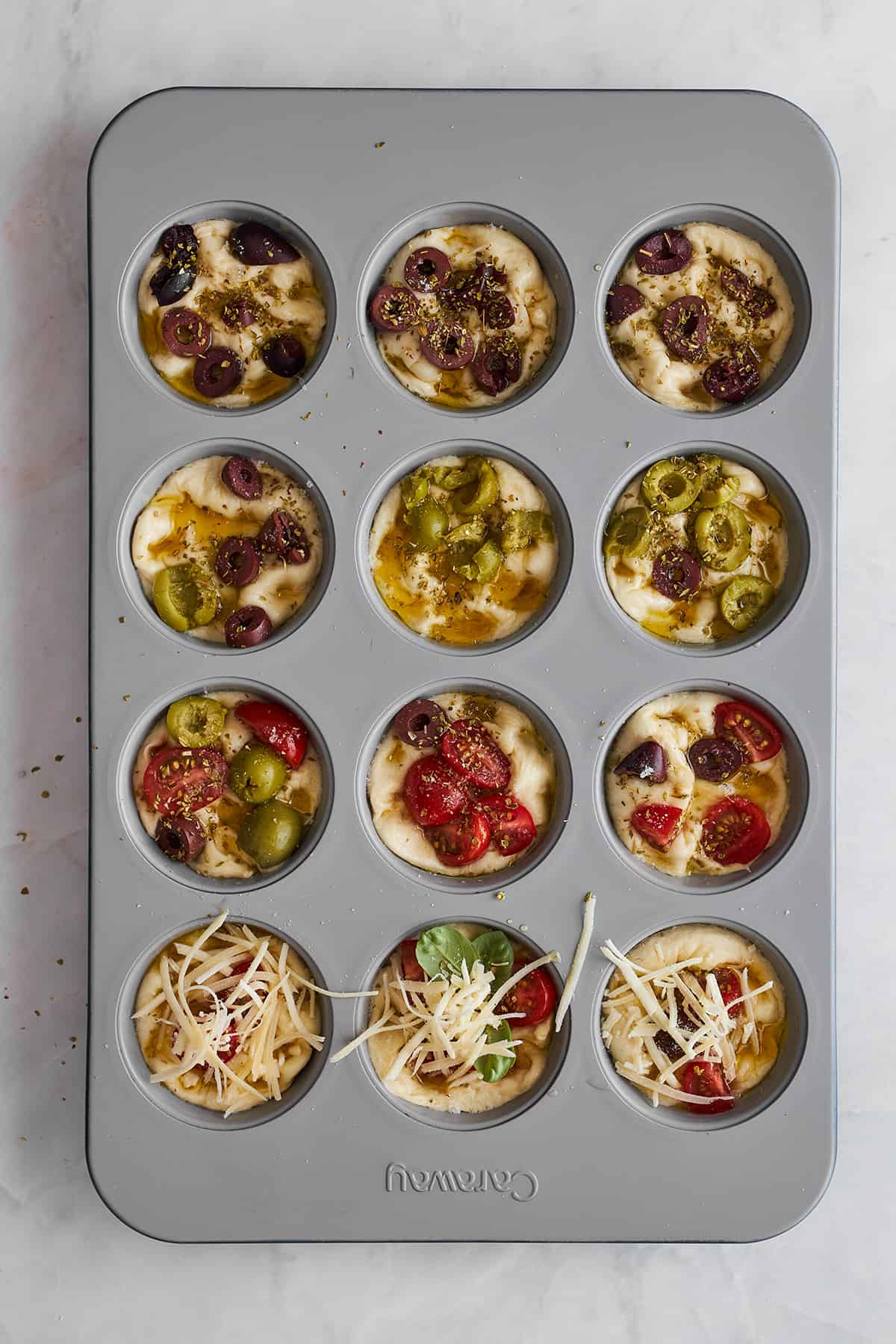 Raw bread focaccia muffins with olives, tomatoes, and cheese in a muffin tin.