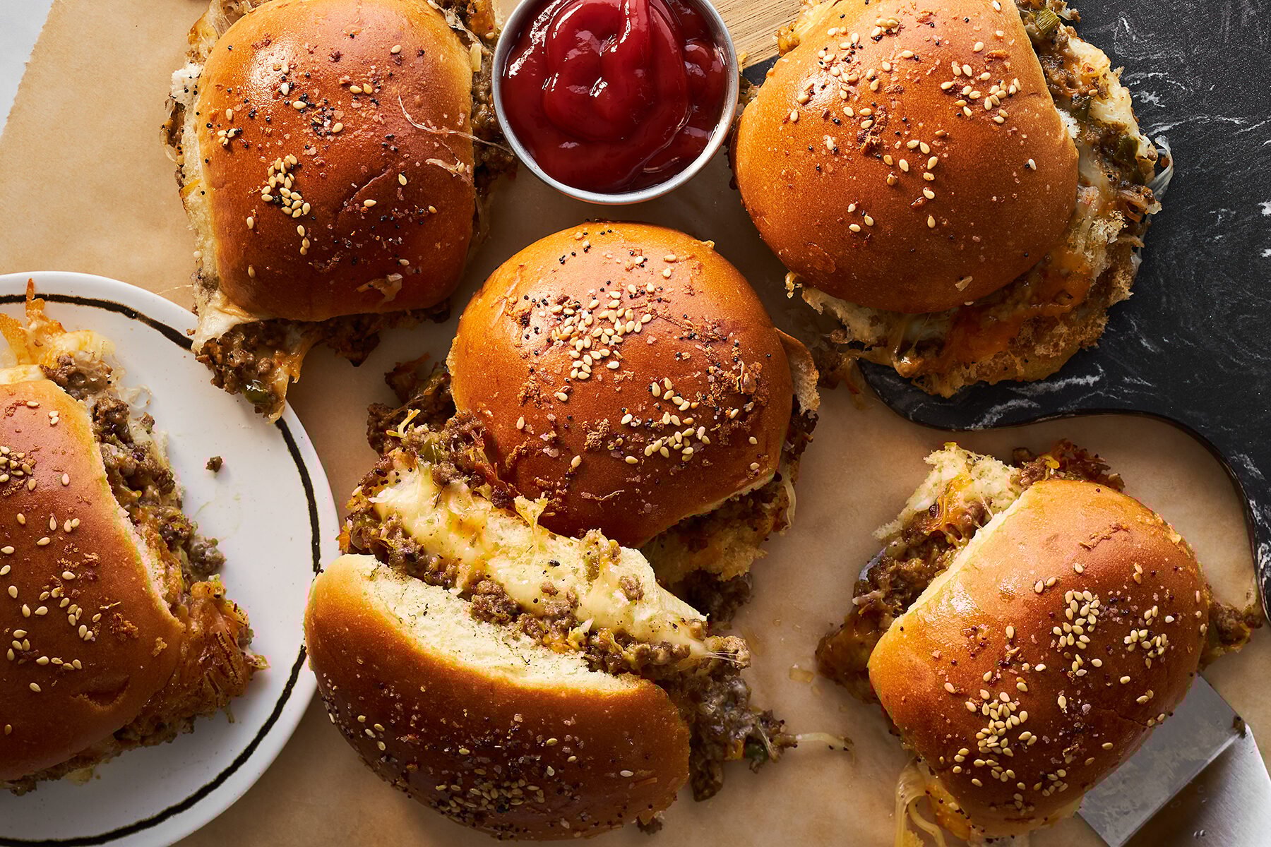 Cheesy ground beef sliders topped with sesame seeds with a small container of ketchup. 