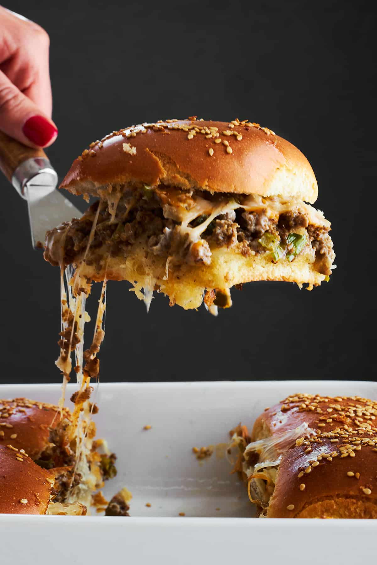 A cheesy ground beef slider being lifted from a baking dish. 