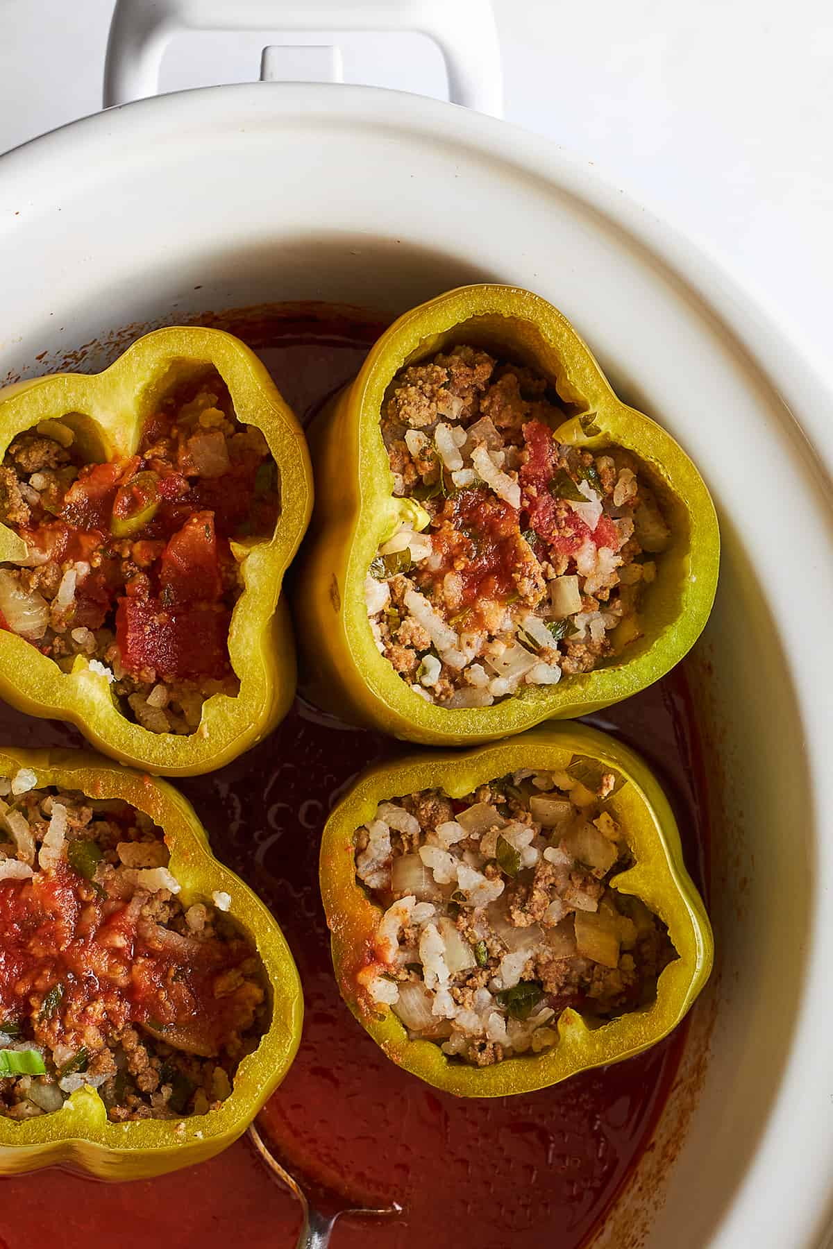Stuffed peppers in a crockpot with a spoon stirring tomato sauce in the bottom. 