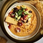 A bowl of spicy feta tip topped with veggies and a cracker.