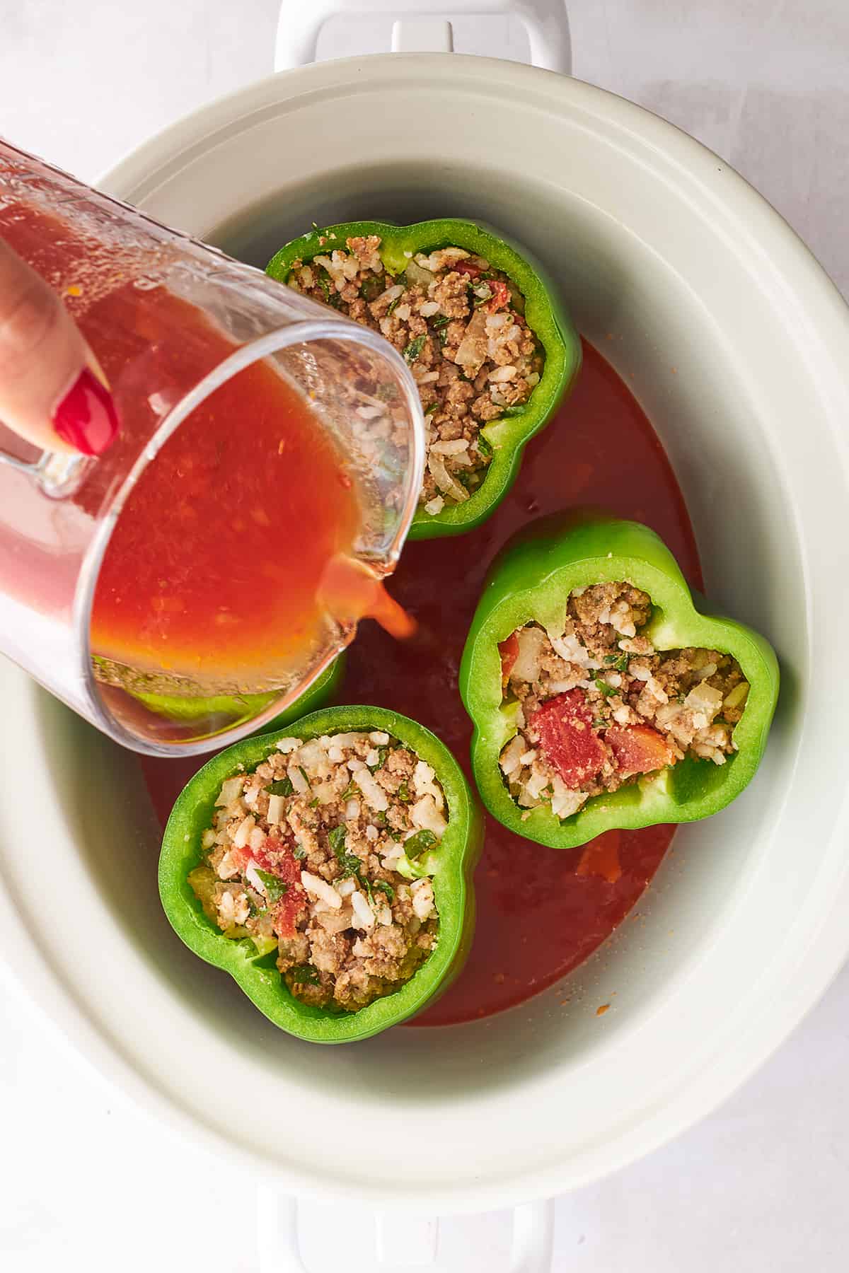 Tomato sauce being poured over stuffed peppers in a crockpot. 