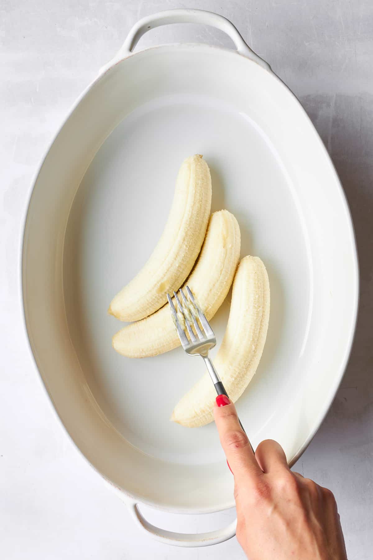 Three bananas in a baking dish being mashed with a fork. 