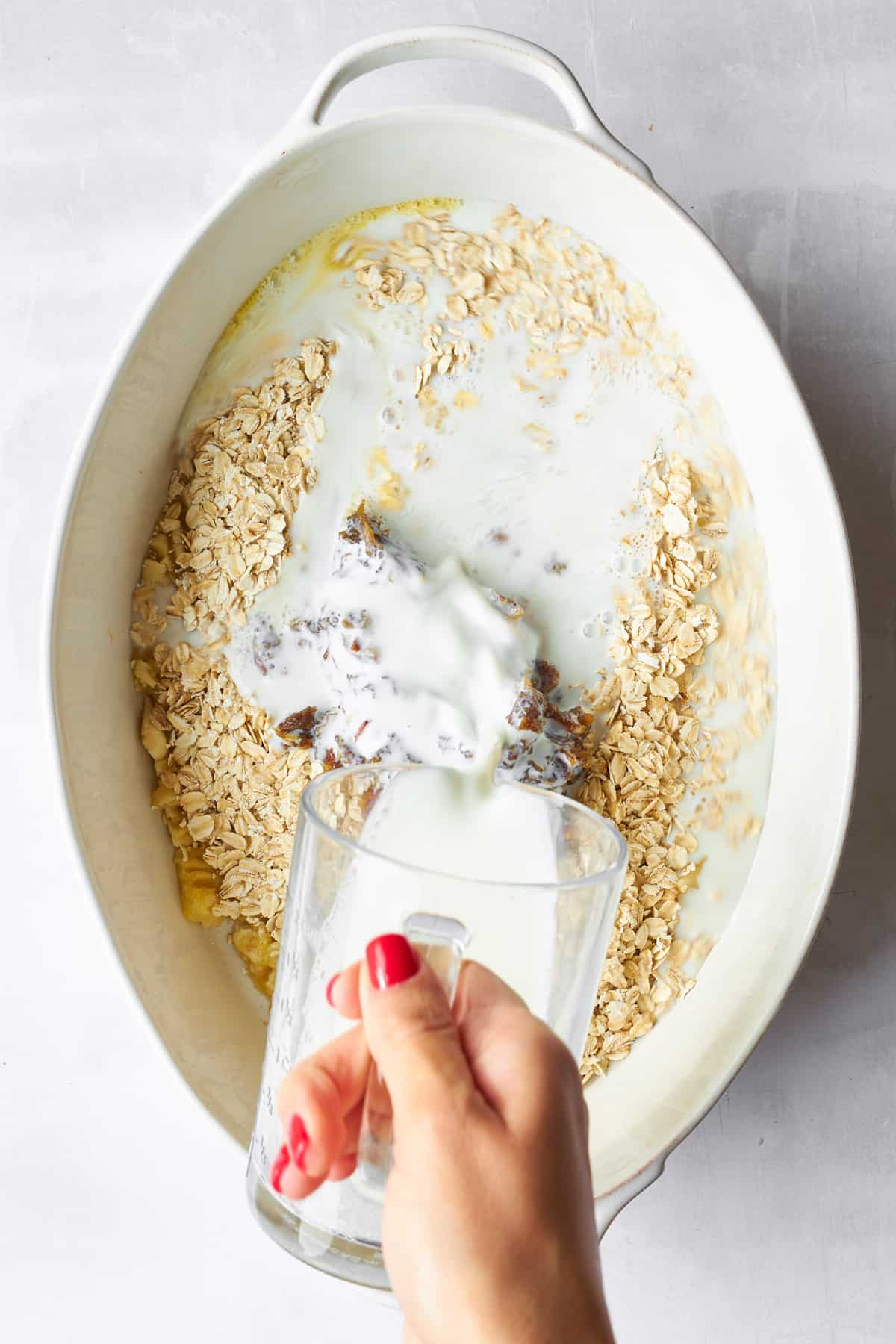 Milk being poured over banana date oatmeal bake ingredients in an oval baking dish. 
