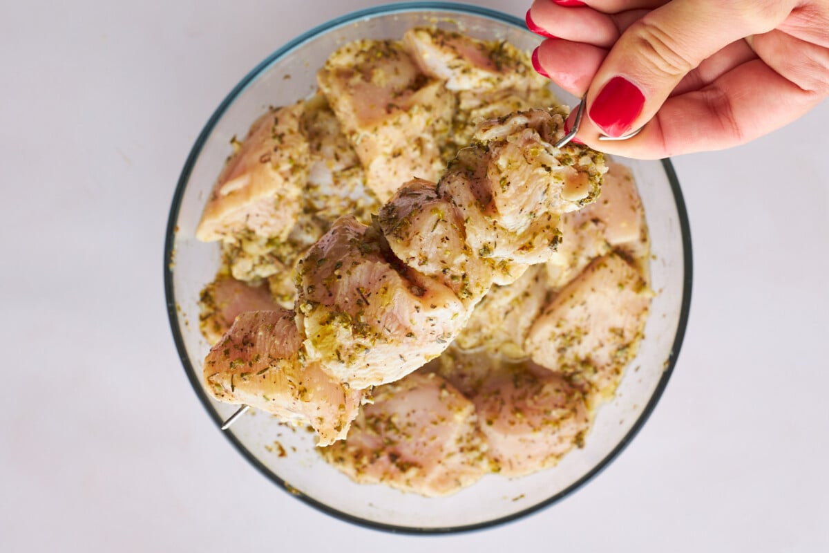 Raw marinaded souvlaki chicken being threaded on skewers.