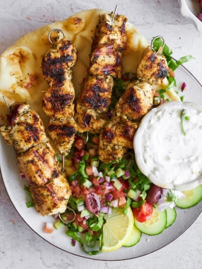 A plate of chicken souvlaki skewers with naan, shirazi salad, and tzatziki.