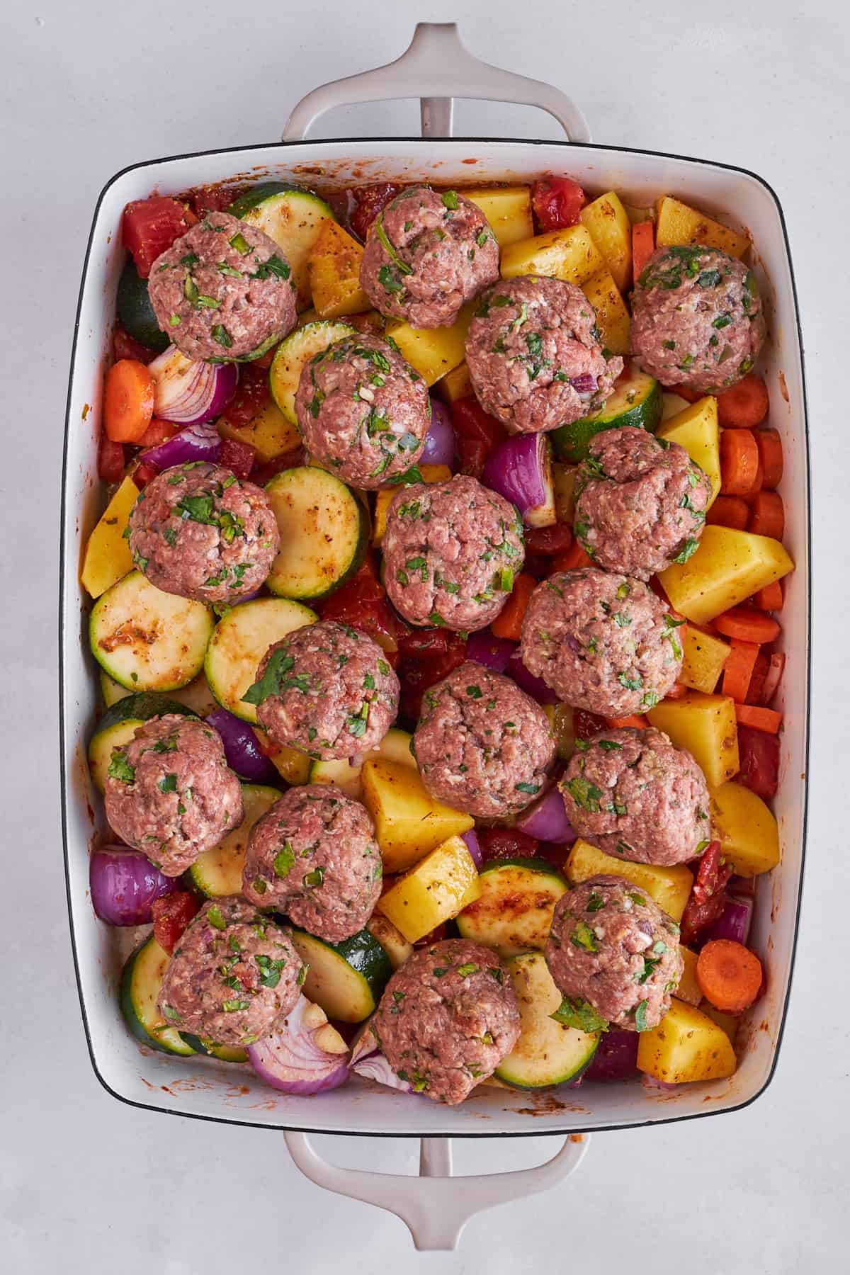 Raw seasoned veggies and raw beef kofta meatballs in a baking dish. 