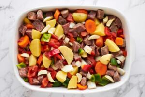 Raw stew meat and sliced veggies in a casserole dish.