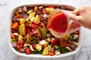 Tomato sauce being poured over beef and veggies in a casserole dish.
