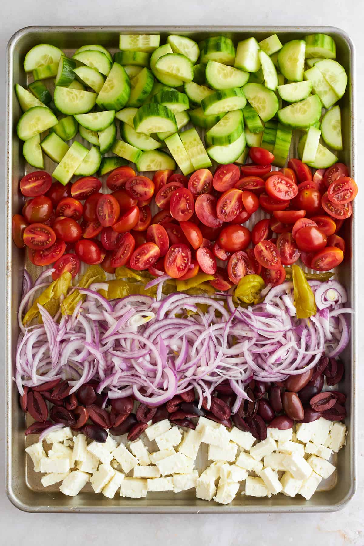 Cucumbers, tomatoes, pepperoncini peppers, red onions, and olives on a baking sheet. 
