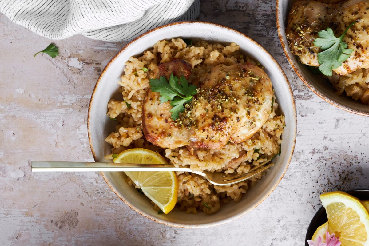 A bowl of Greek chicken and rice with a fork with a second bowl of chicken and rice and two lemon wedges in the background. 