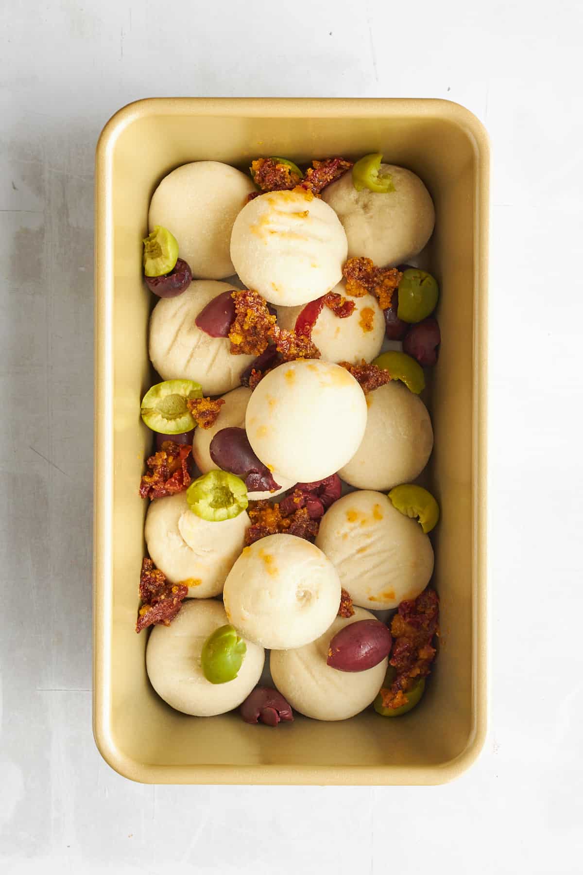 Raw bread rolls, olives, and sun-dried tomatoes in a loaf pan. 