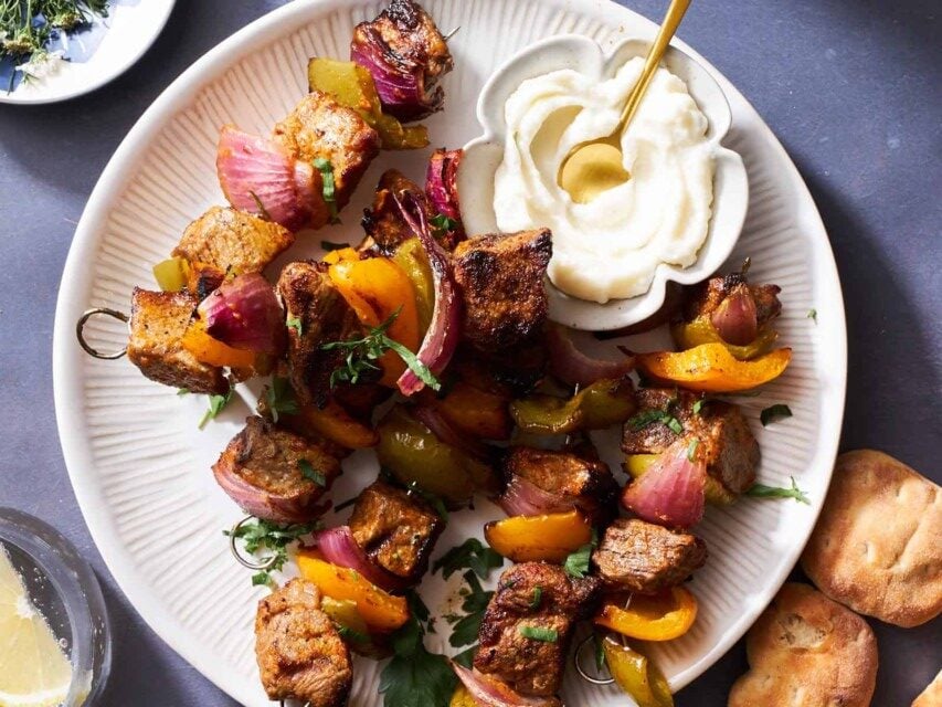 A plate of grilled steak skewers with onions and peppers and a small bowl of toum on top.