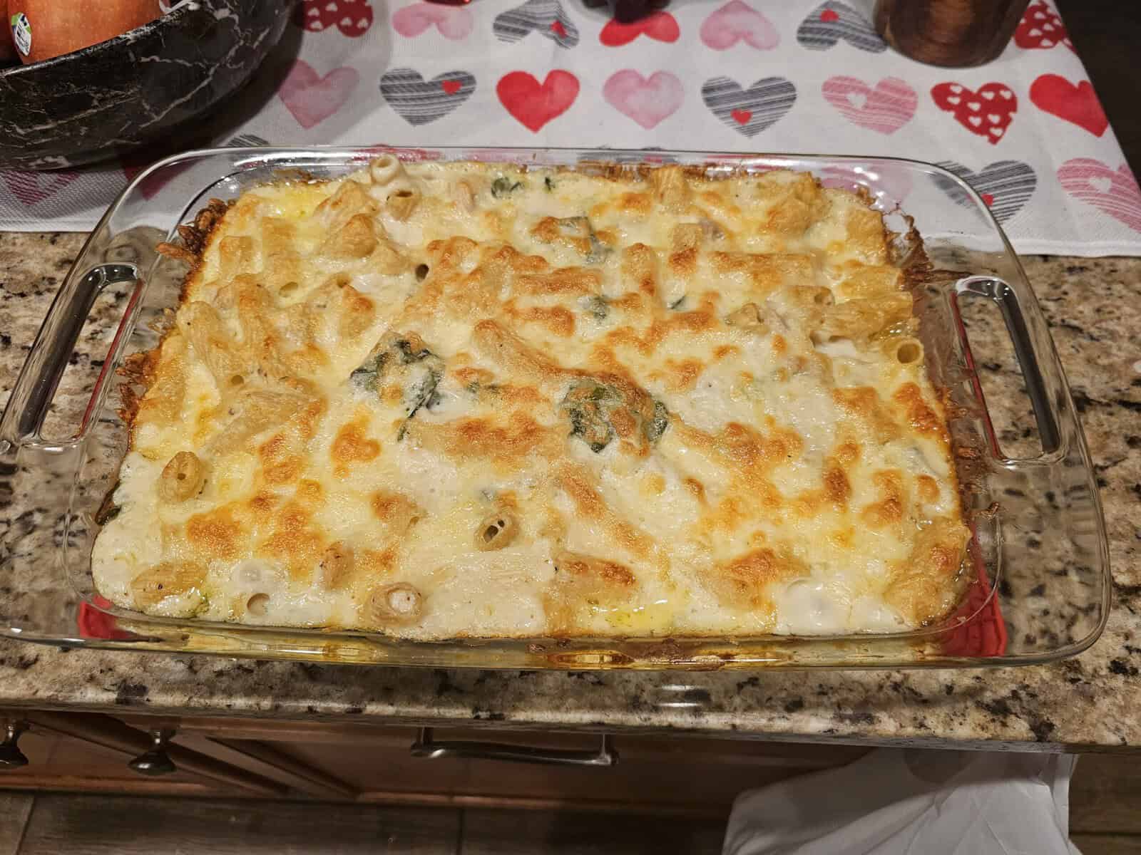 A glass baking dish filled with creamy baked pasta, topped with melted cheese and bits of spinach. The dish sits on a kitchen countertop, partially covering a heart-patterned tablecloth.