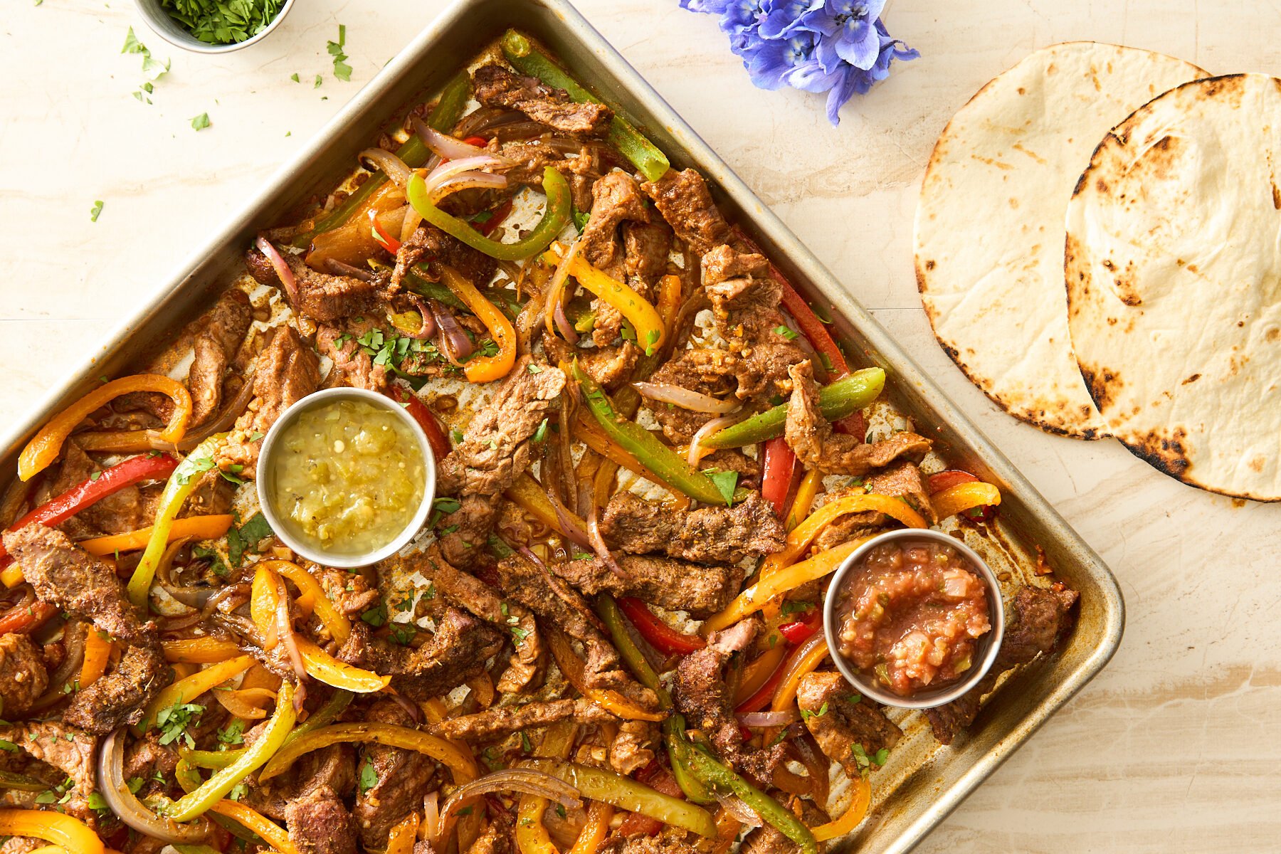 Sheet pan steak fajitas on a baking sheet with salsa verde and red salsa next to two tortillas. 