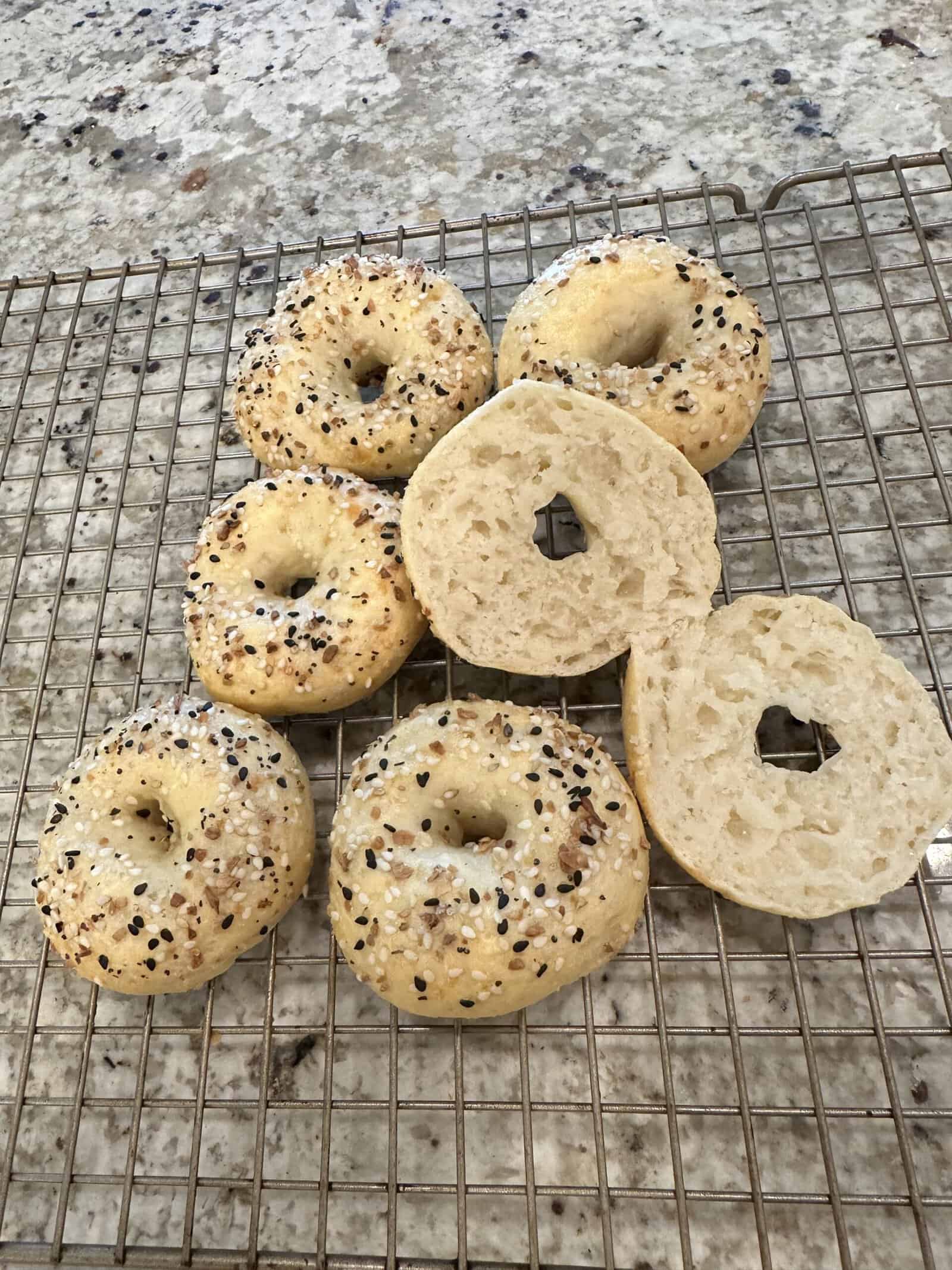 Seven homemade bagels with everything seasoning on a cooling rack. One bagel is sliced in half, showing a soft interior. The rack sits on a speckled countertop.