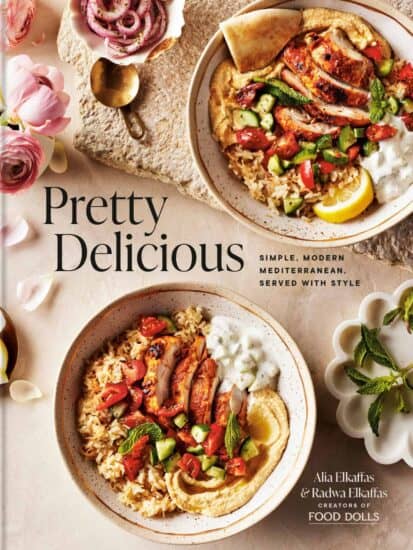 A cookbook titled Pretty Delicious featuring Mediterranean dishes. The cover shows two bowls with rice, grilled vegetables, falafel, and pita, garnished with lemon and herbs. Pink flowers and a small dish with yogurt are nearby.