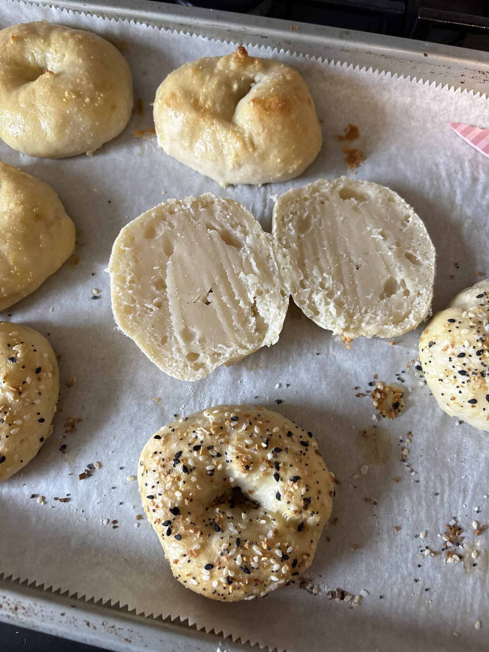 Six homemade bagels on a baking tray lined with parchment paper. One bagel is sliced open, showing a dense, soft interior. The others are topped with various seasonings, including everything bagel seasoning.