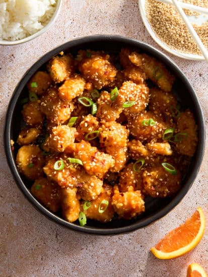 A bowl of cooked orange chicken topped with sesame seeds and scallions.