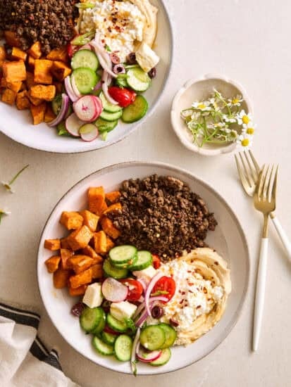 Two ground beef bowls with beef, roasted sweet potatoes, fresh veggies, feta, hummus, and cottage cheese.