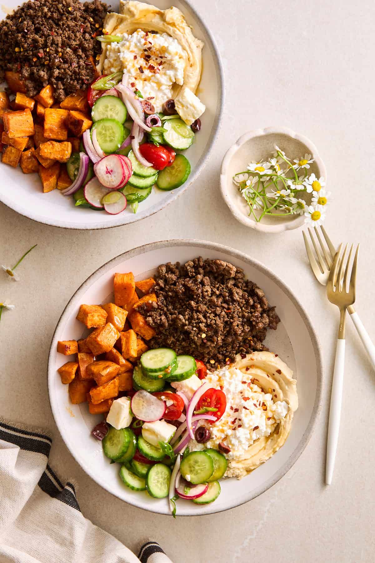 Two ground beef bowls with beef, roasted sweet potatoes, fresh veggies, feta, hummus, and cottage cheese.