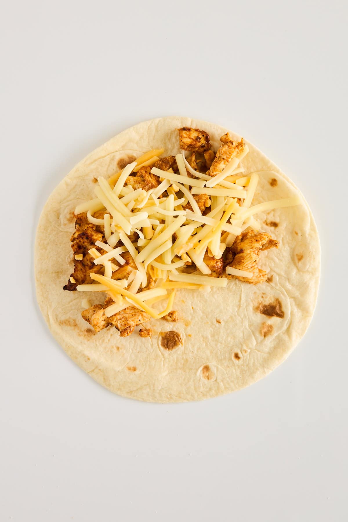 A flour tortilla on a white surface topped with seasoned cooked chicken pieces and shredded cheese, ready to be folded or wrapped.