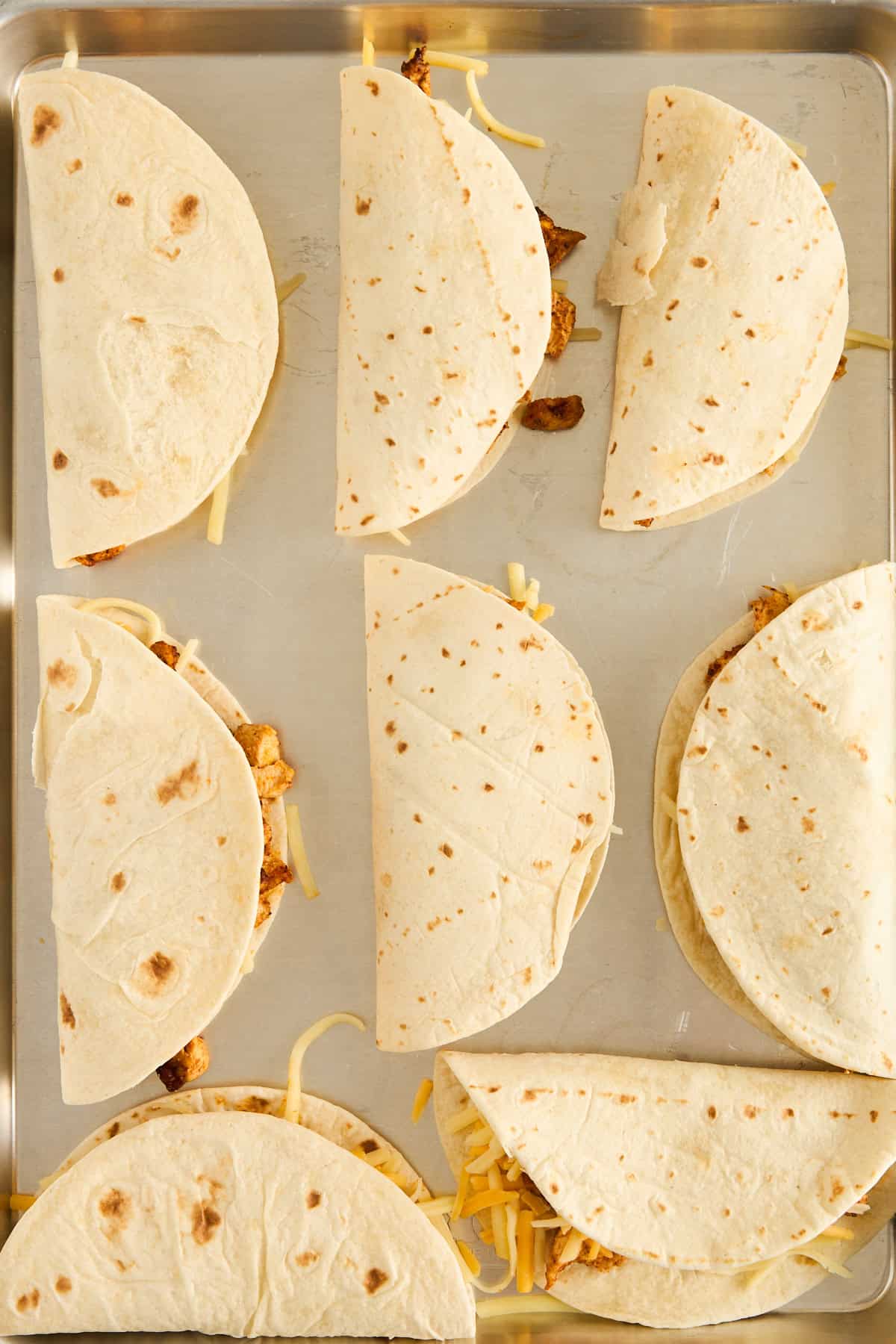 Eight flour tortillas filled with cheese and a meat mixture are arranged in neat rows on a metal baking sheet, ready to be baked.