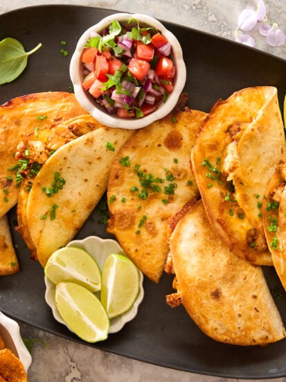 A black platter with six crispy tacos garnished with chopped chives, served with lime wedges and a small bowl of fresh tomato salsa. Tortilla chips sit in a dish nearby.