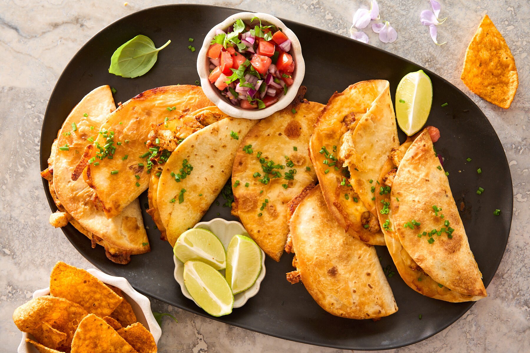A black platter with six crispy tacos garnished with chopped chives, served with lime wedges and a small bowl of fresh tomato salsa. Tortilla chips sit in a dish nearby.