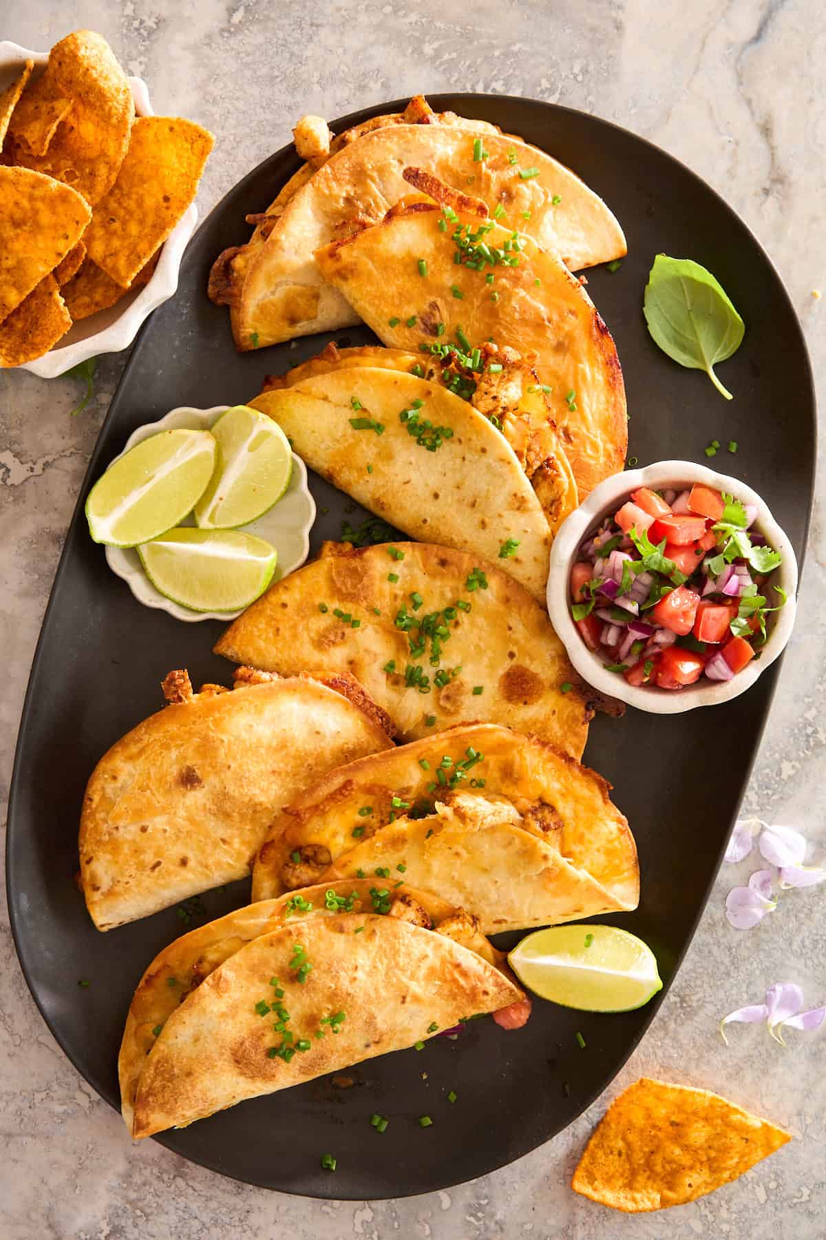 A black platter with six crispy tacos garnished with chopped chives, served with lime wedges and salsa. 