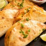 A close-up of crispy tacos filled with melted cheese, garnished with chopped chives, and served with lime wedges and a small bowl of salsa on the side.