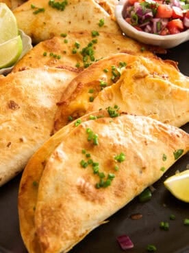 A close-up of crispy tacos filled with melted cheese, garnished with chopped chives, and served with lime wedges and a small bowl of salsa on the side.