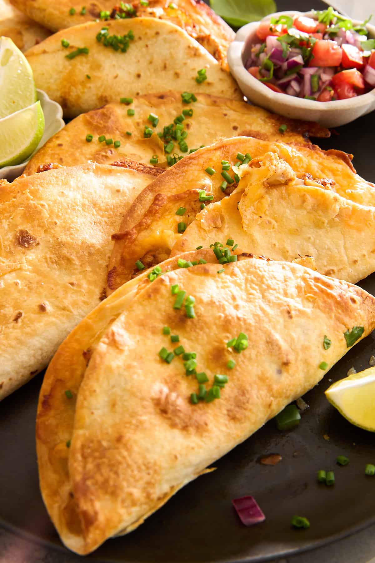 A close-up of crispy tacos filled with melted cheese, garnished with chopped chives, and served with lime wedges and a small bowl of salsa on the side.