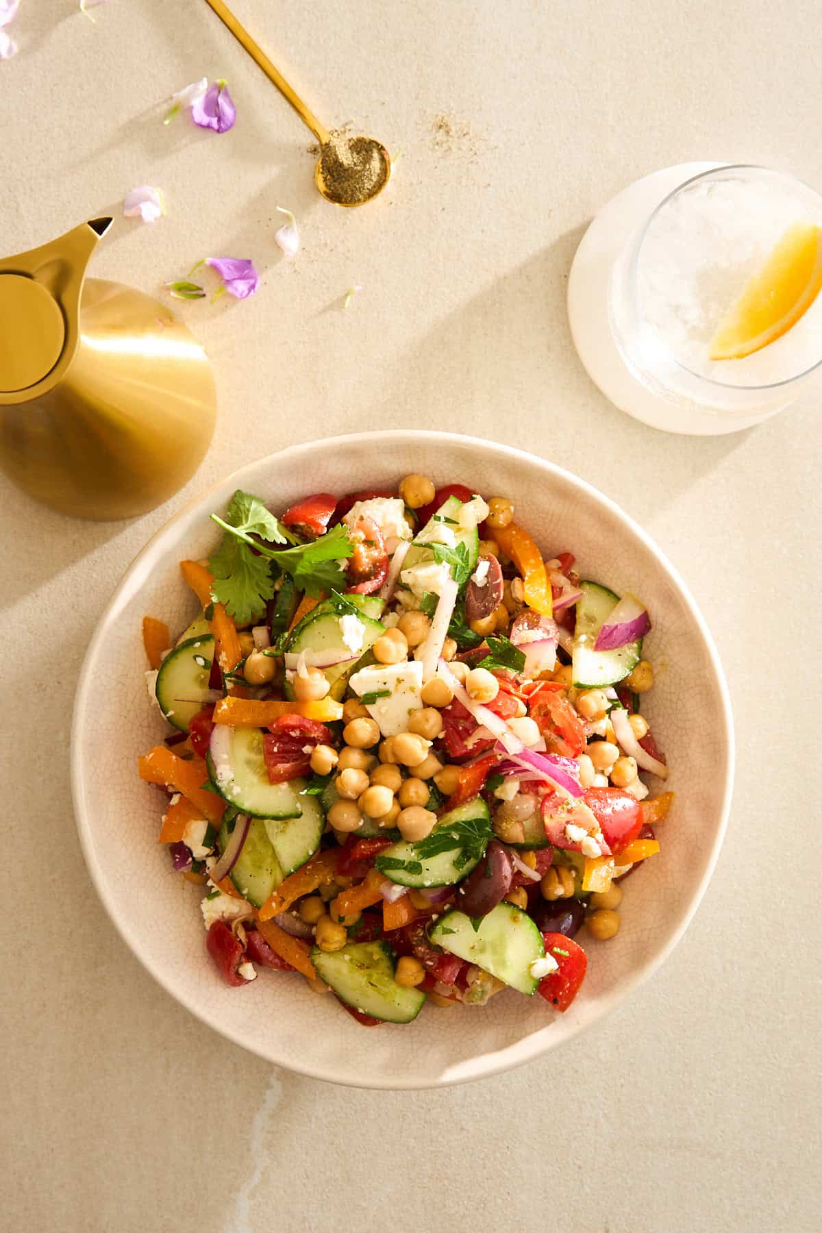 A bowl of colorful chickpea salad with cucumbers, tomatoes, olives, red onion, and herbs on a light surface, next to a gold pitcher, a spoon with spices, and a glass with ice and a lemon twist.
