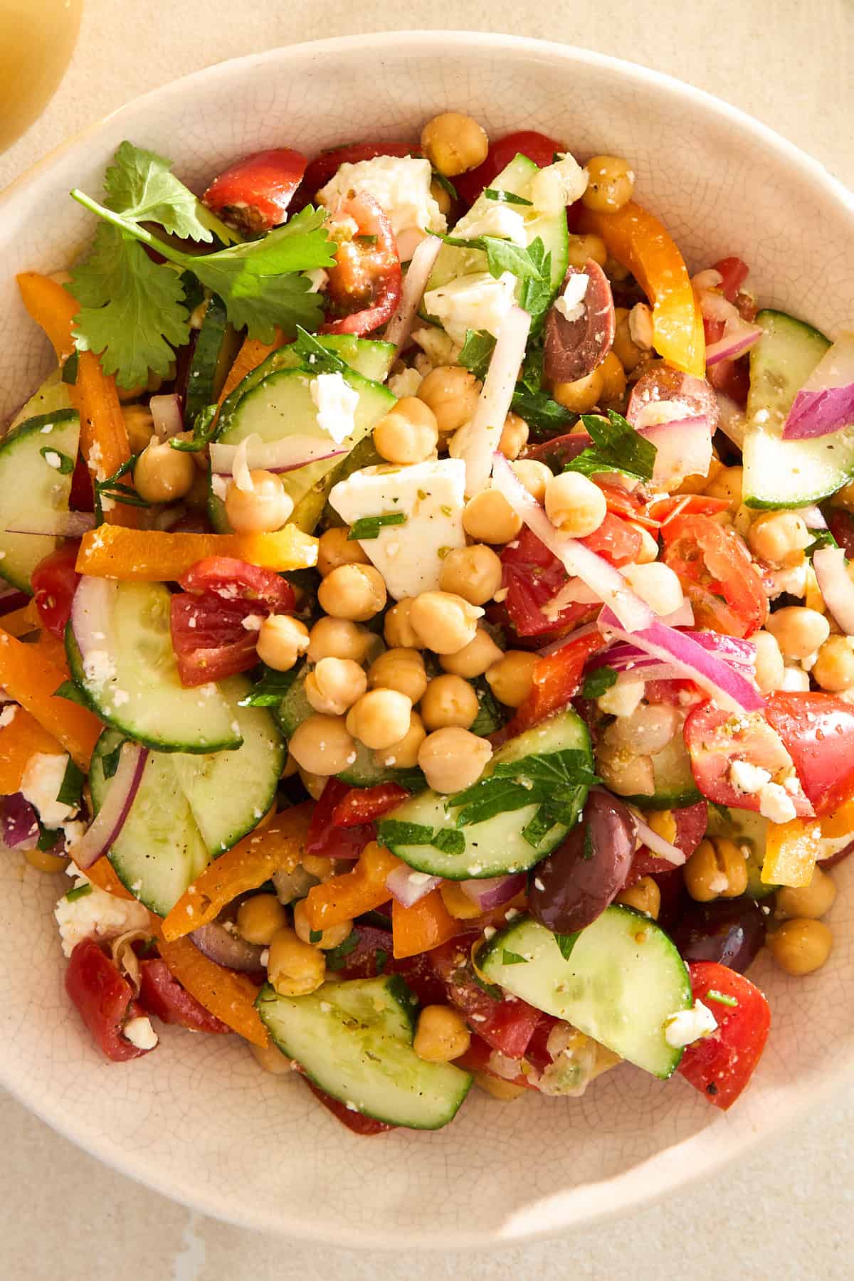 A bowl of colorful salad with chickpeas, sliced cucumbers, cherry tomatoes, red onion, yellow bell pepper, olives, feta cheese, and fresh herbs.
