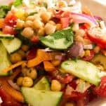A close-up of a fresh salad with sliced cucumbers, tomatoes, chickpeas, red onions, carrots, olives, and herbs in a white bowl.