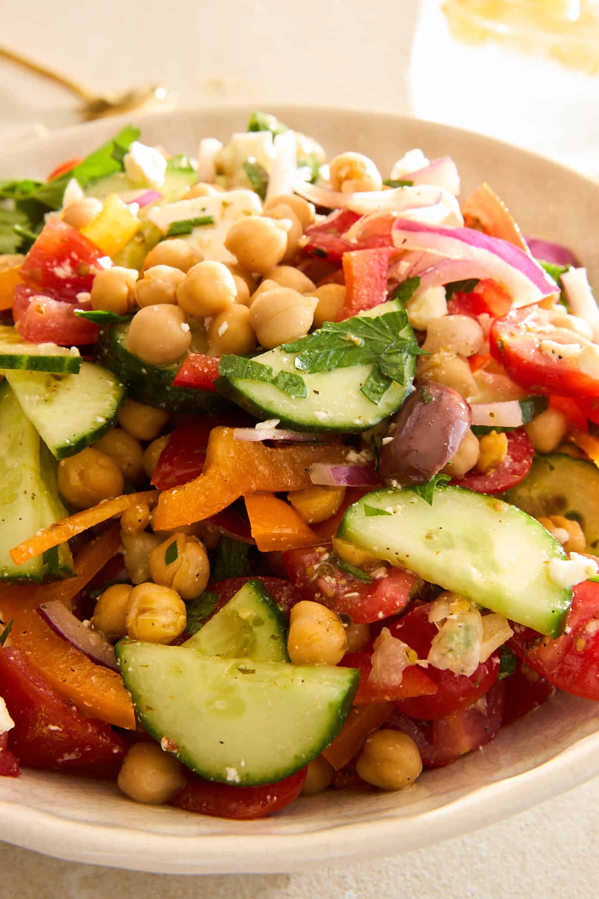 A close-up of a fresh salad with sliced cucumbers, tomatoes, chickpeas, red onions, carrots, olives, and herbs in a white bowl.