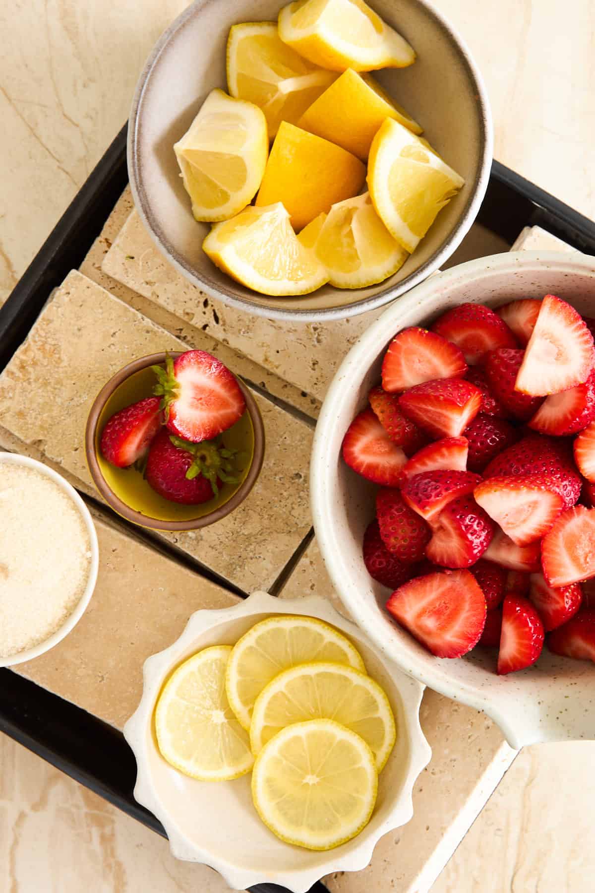 Bowls containing sliced lemons, quartered strawberries, whole strawberries, granulated sugar, and lemon slices.