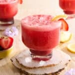 A glass of pink strawberry lemonade with ice, garnished with a lemon slice and strawberry, sits on stone coasters. Two similar drinks and small bowls of strawberries and lemon slices are in the background.