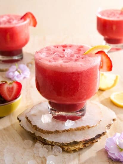 A glass of pink strawberry lemonade with ice, garnished with a lemon slice and strawberry, sits on stone coasters. Two similar drinks and small bowls of strawberries and lemon slices are in the background.